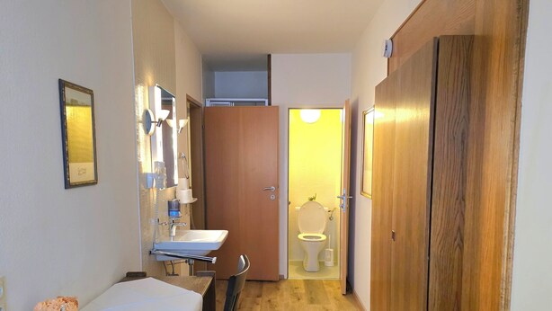 A hotel room with a wooden desk, a chair, a bed, a small table with a vase, and a mirror. There is a wooden wardrobe and a door leading to another room. - Zum Goldenen Kreuz (Eferding)