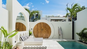 Un área de piscina exterior moderna con diseño blanco y de madera, que incluye un sofá, jardineras y una puerta de madera.