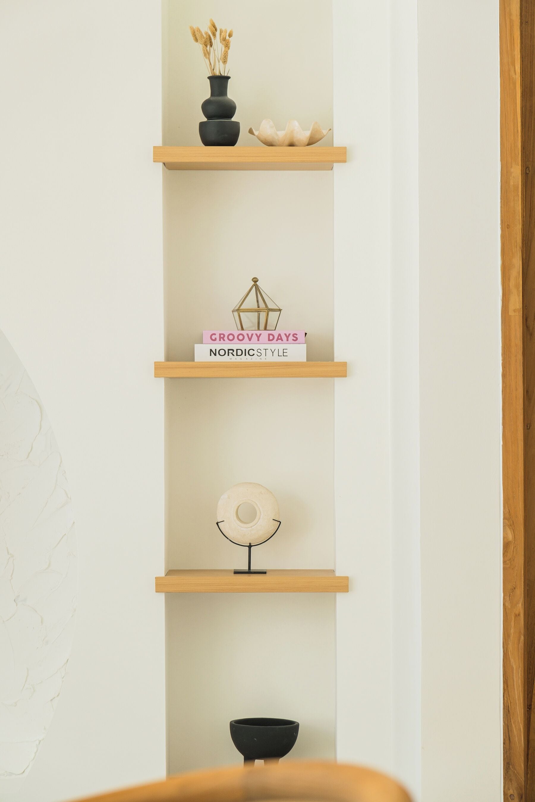A wall-mounted shelf with decorative items including a vase with dried plants, a bowl, and a book titled 'Groovy Days Nordic Style'.
