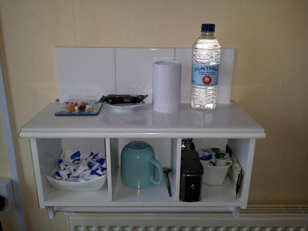 A white minibar with a bottle of water, a roll of toilet paper, a plate with snacks, a mug, and a coffee maker.