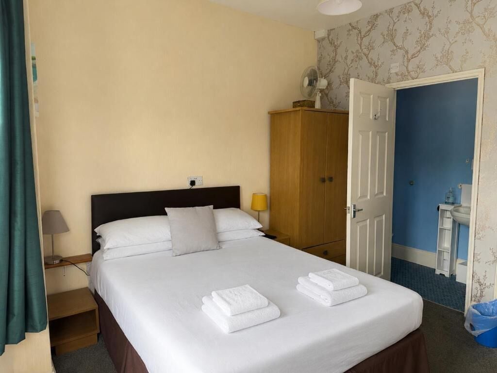 Ein Hotelzimmer mit einem Bett, einem Holzschrank, einem Nachttisch mit Lampe und einem durch eine offene Tür einsehbaren Badezimmer.