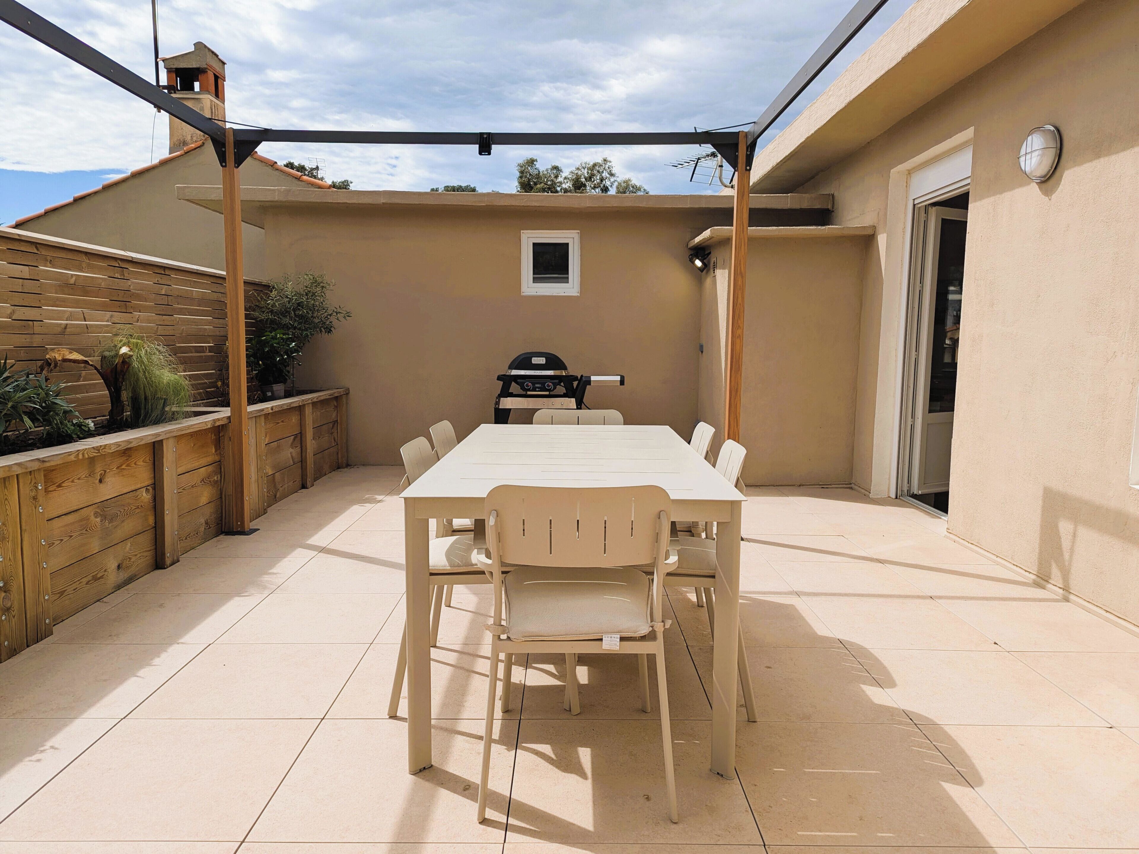 Een overdekt terras met een eettafel en stoelen, een barbecue en een houten plantenbak met planten.