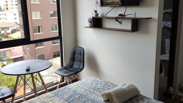 Ein Schlafzimmer mit einem Bett, einem Fernsehgerät an der Wand, einem kleinen Tisch, einem Stuhl und einem Fenster mit Blick auf Gebäude.