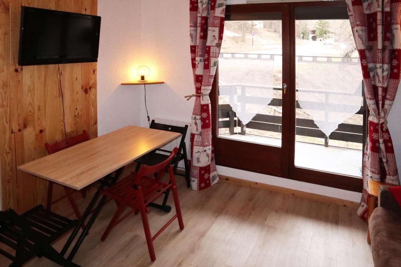 Une pièce avec une table en bois, des chaises rouges, un téléviseur fixé au mur, une lampe et une vue sur l’extérieur à travers une grande fenêtre ornée de rideaux à motifs.