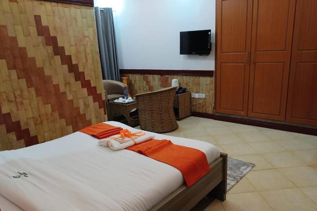 A hotel room with a bed, a television on the wall, a wooden wardrobe, and a small table with a chair.