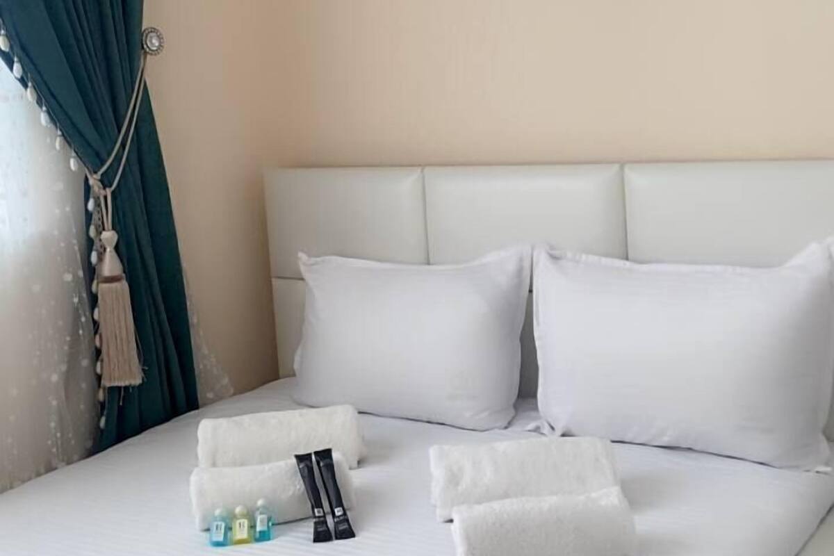 A neatly made bed with white linens, a painting of flowers on the wall, and a blue curtain.