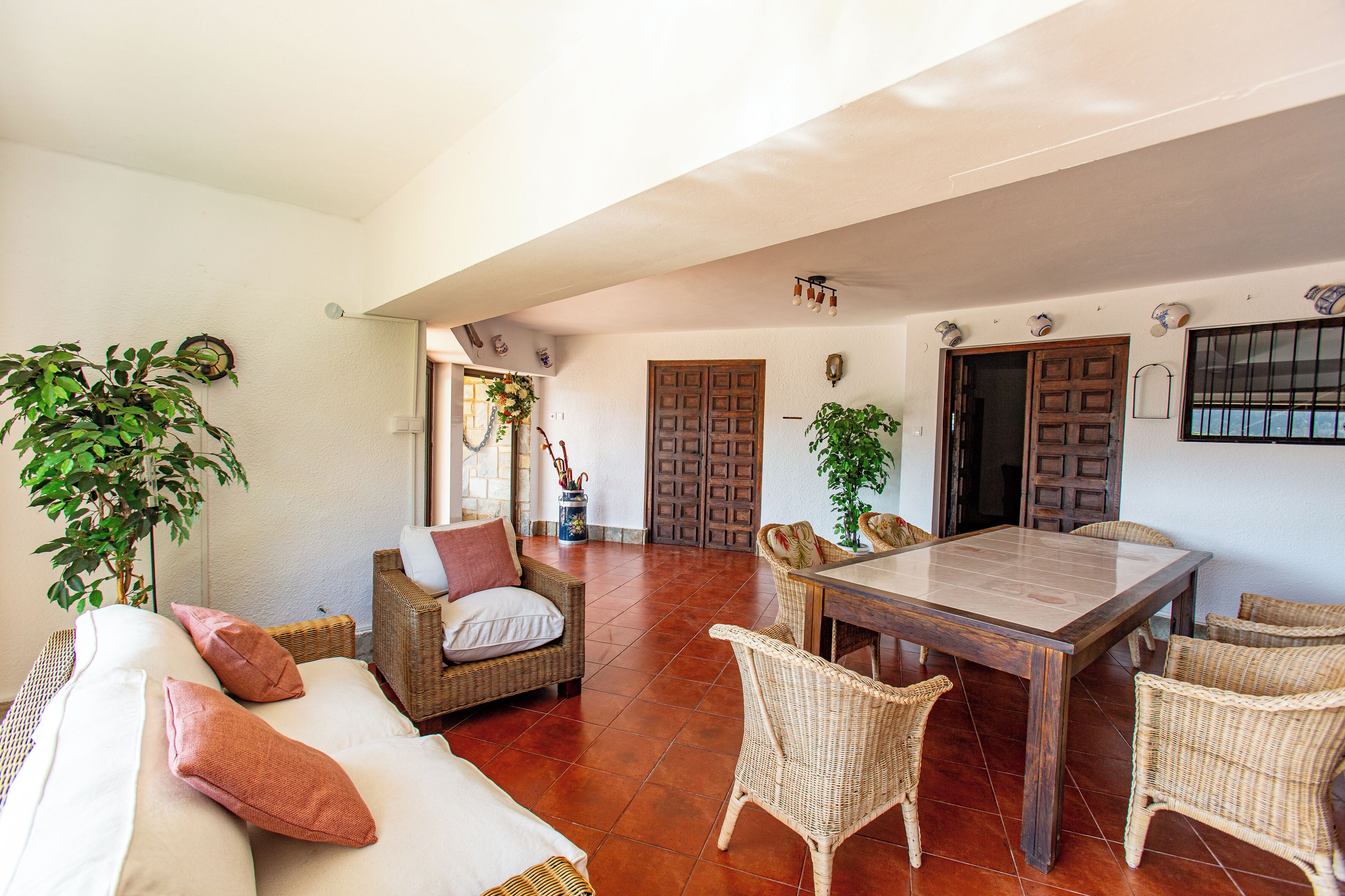 A spacious living area with a large dining table, wicker furniture, and a white sofa with cushions.