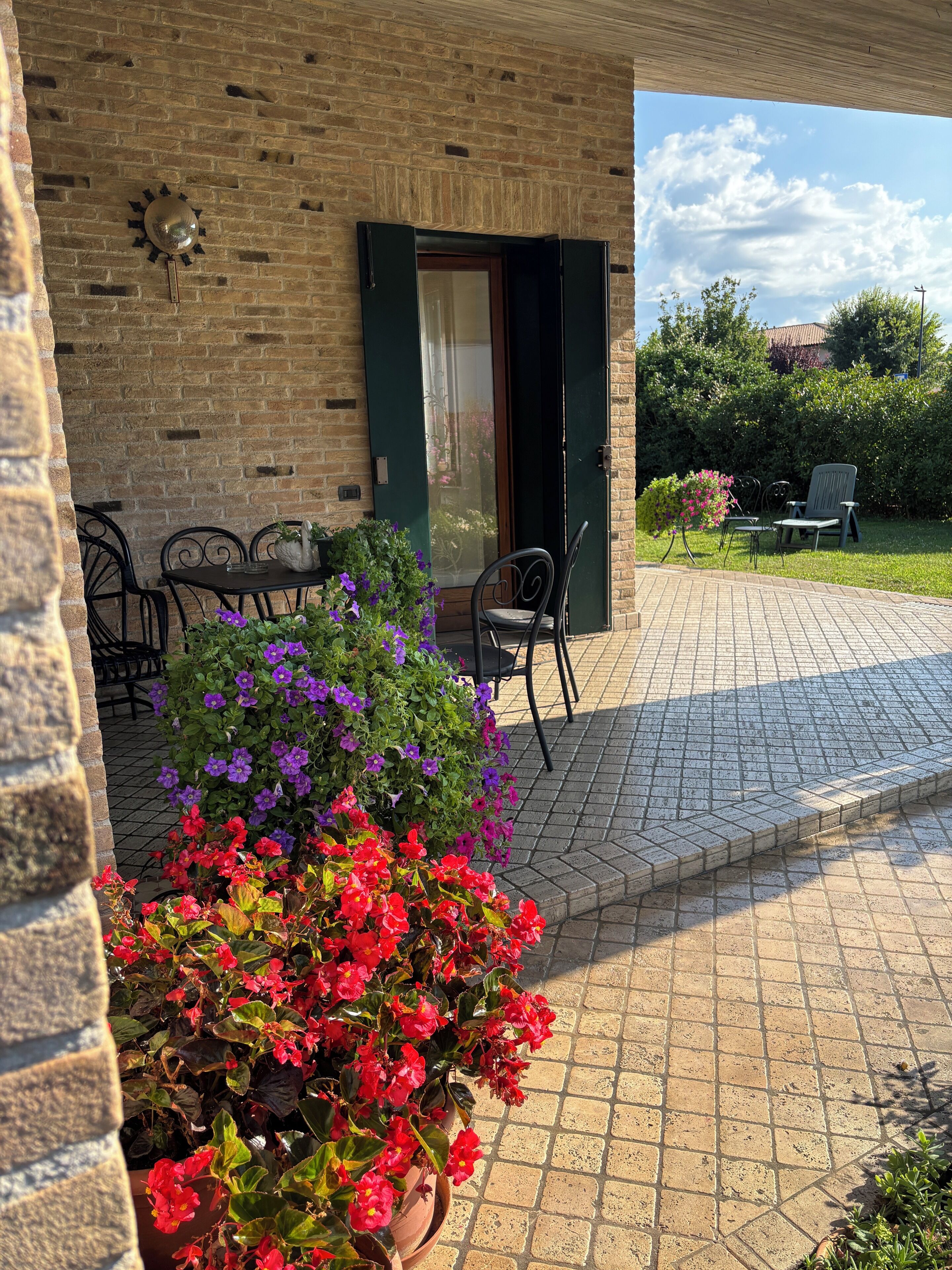 A brick building with a patio area featuring a table and chairs, surrounded by potted plants with red and purple flowers.