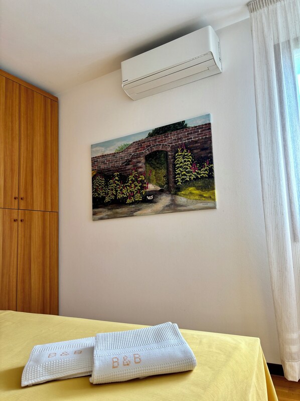 A room with a yellow table, a painting of a bridge, an air conditioning unit, and a wooden wardrobe. - B&B Alla Suite (Pieve di Soligo)
