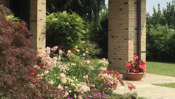 A brick building with a covered entrance, surrounded by lush greenery and flowering plants. - B&B Alla Suite (Pieve di Soligo)