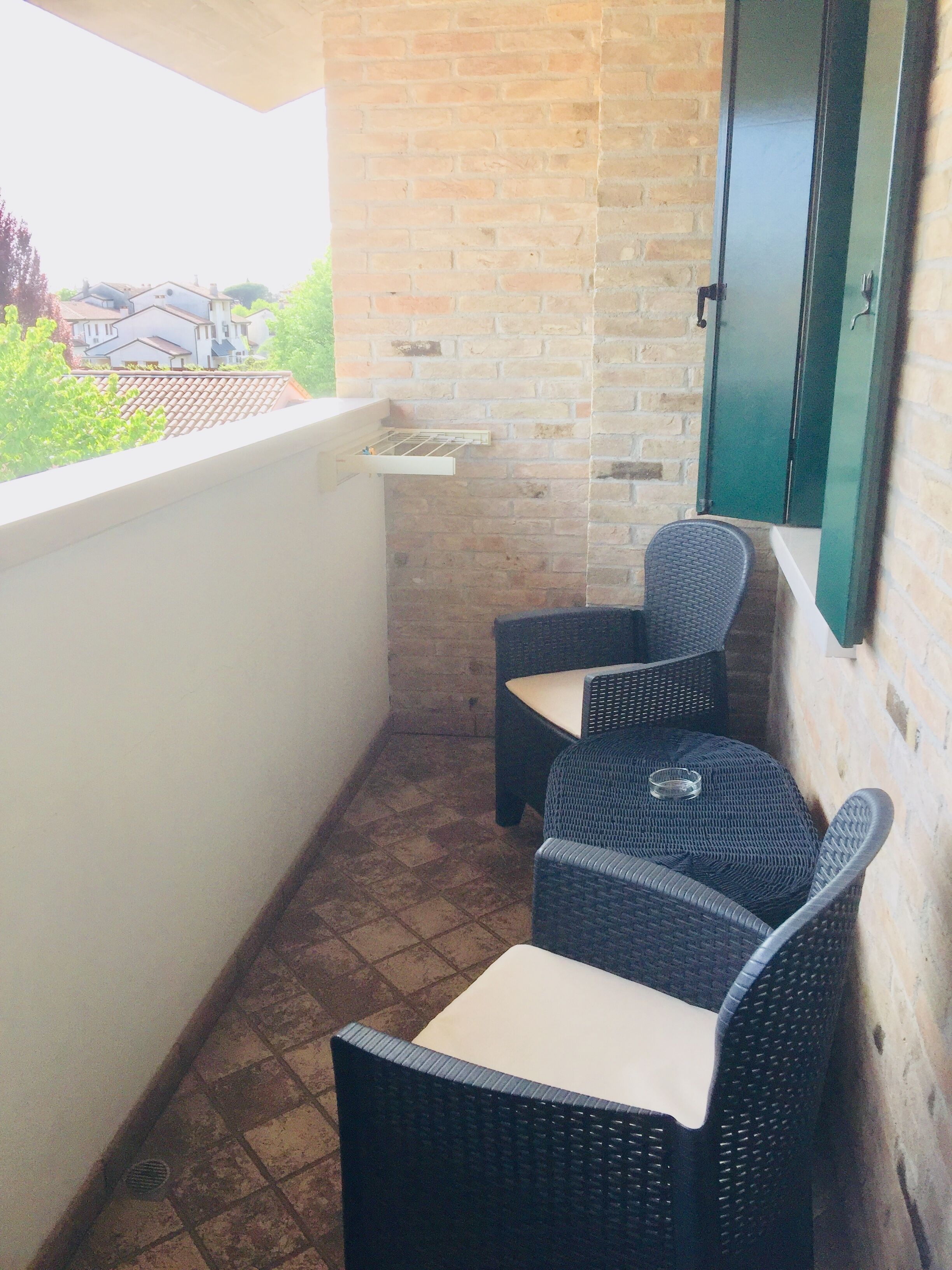 Un pequeño balcón con dos sillones de mimbre y una mesa, un muro de ladrillo y vistas a casas al fondo.