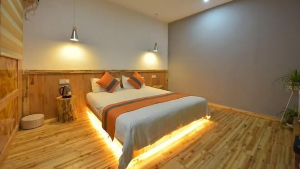 Ein modernes Schlafzimmer mit Holzboden, einem Bett mit orange-weißer Bettwäsche, zwei Pendelleuchten und einem kleinen runden Tisch mit einer Kaffeemaschine.
