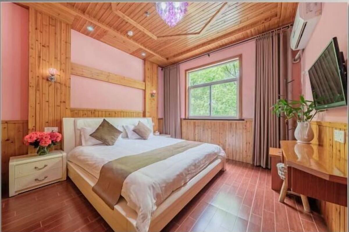 A bedroom with a wooden ceiling, a large bed, a bedside table with a vase of flowers, and a window with curtains.