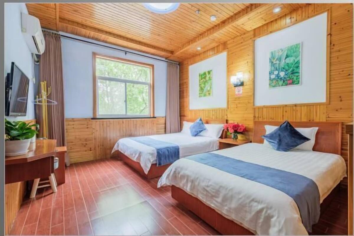 A room with wooden paneling, two beds with blue and white bedding, a desk with a computer, a window with curtains, and artwork on the walls.