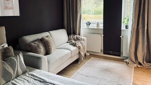 Un salón moderno con un sofá gris, una lámpara de pie tejida y vistas al exterior con mucha vegetación a través de una ventana.