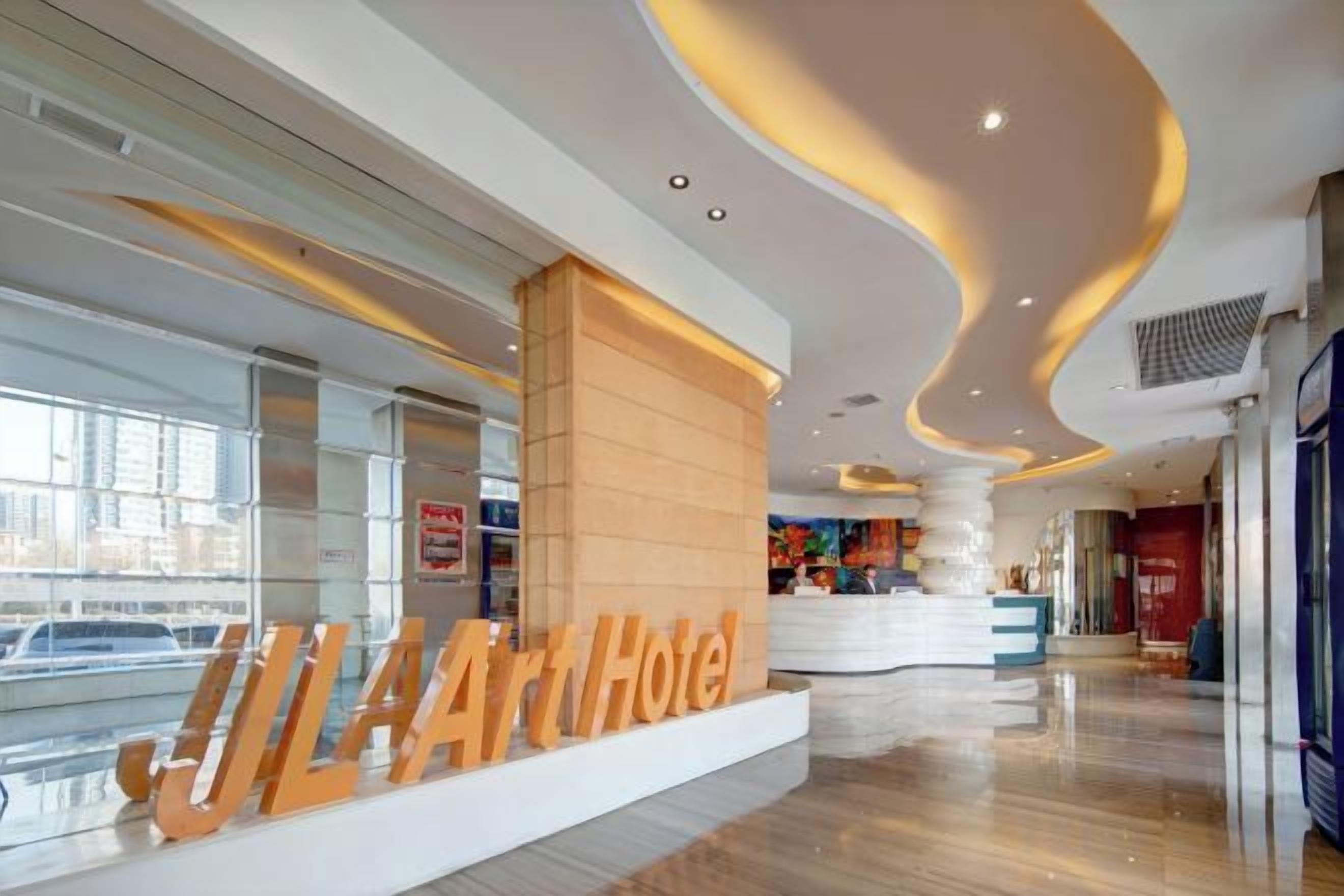 A hotel lobby with a reception desk, a large sign reading 'JLA Art Hotel,' and a modern ceiling design.