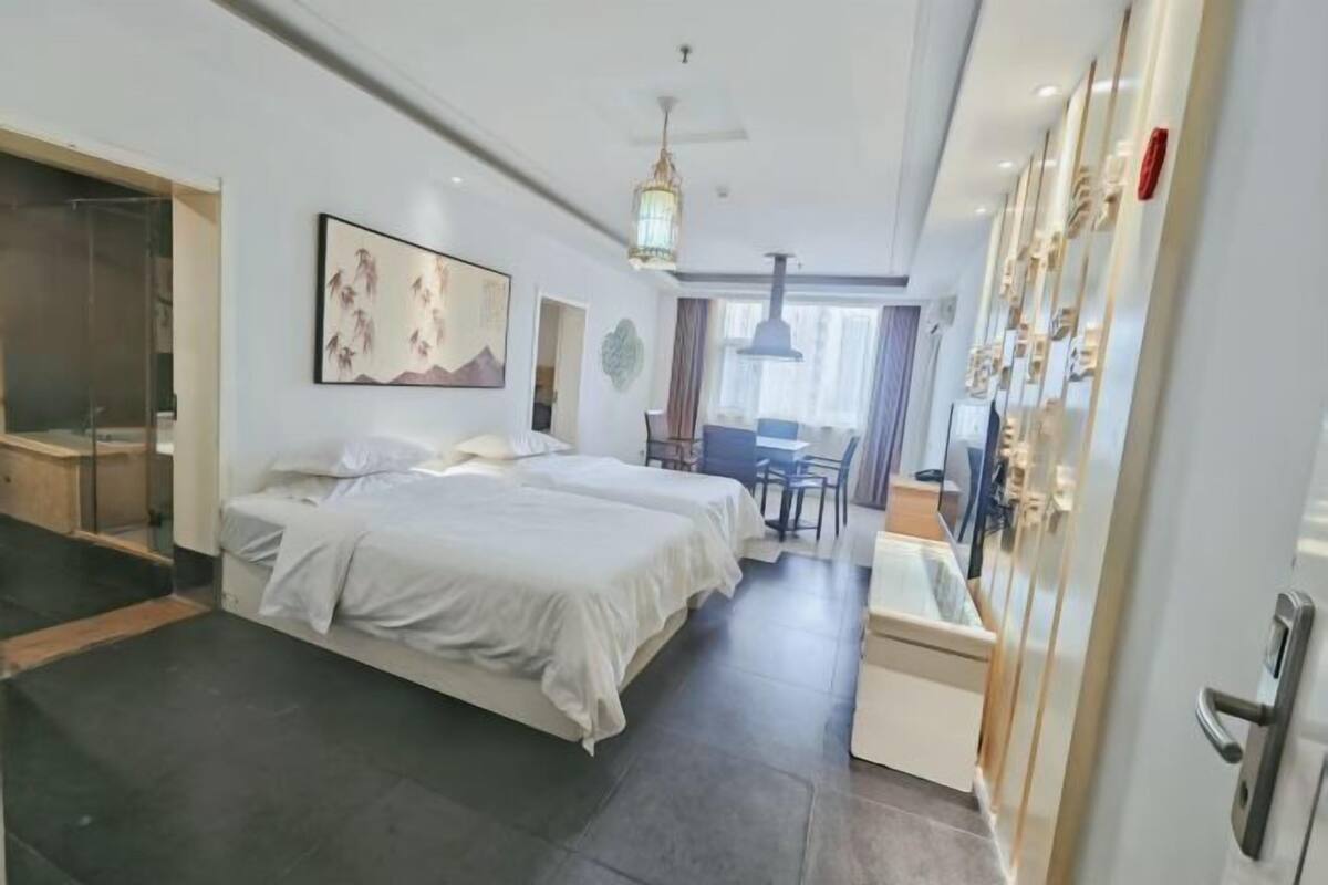 Habitación de hotel con dos camas, un escritorio y una silla. Cuenta con un ventanal con cortinas, un ventilador de techo y una obra de arte colgada en la pared.