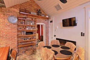 Tunnelmallinen olohuone, jossa on tiilistä tehty takka, seinälle asennettu televisio sekä ruokailutila, jossa on pyöreä pöytä ja pehmeät tuolit.