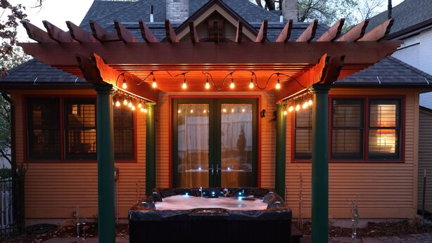 Een hottub onder een houten pergola met lichtjes, omgeven door een kiezelpad en een huis met een puntige dakkapel.