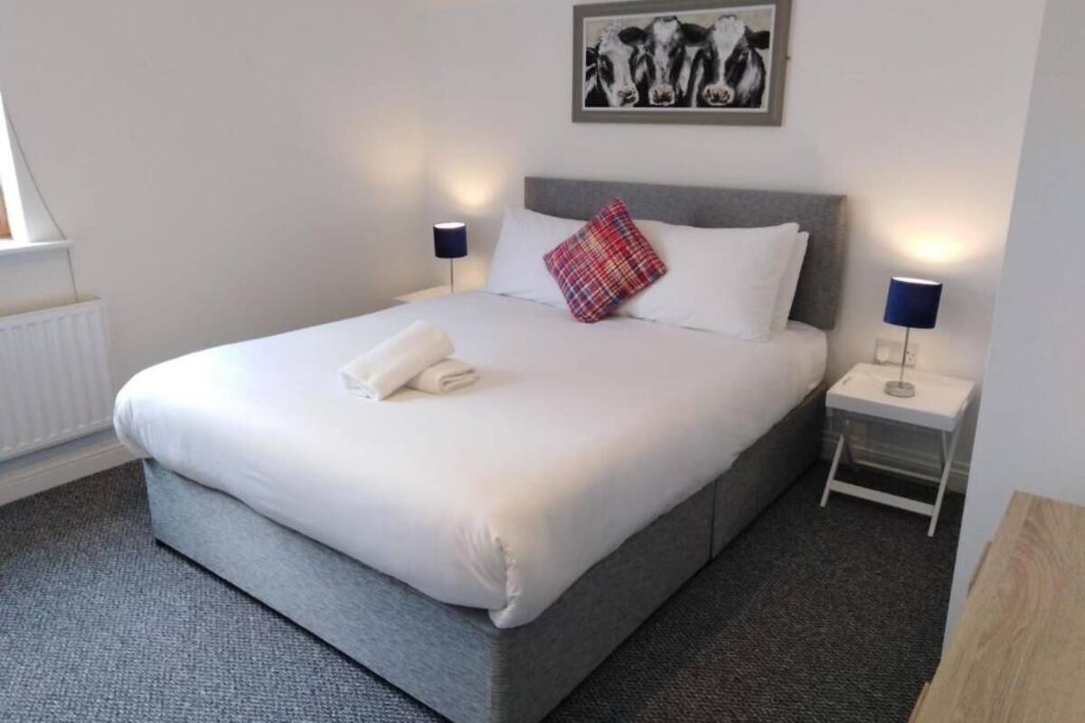 Een net opgemaakt bed met witte lakens en een rood-blauwe ruitkussen, een grijs hoofdbord en twee bedlampen op witte nachtkastjes.