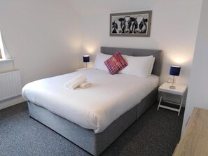 A neatly made bed with white linens and a red and blue plaid pillow, a gray headboard, and two bedside lamps on white nightstands. - Burren Court Lodge (Ballyvaughan)