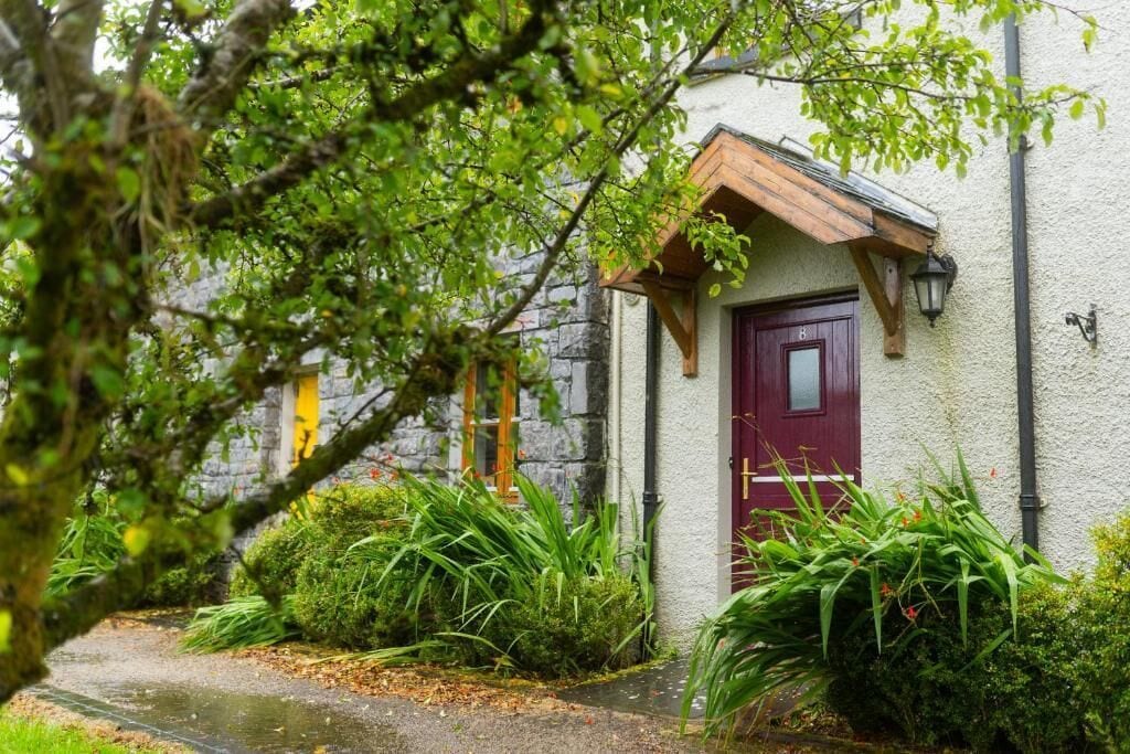 A rustic building with a red door, surrounded by lush greenery and a stone wall.
