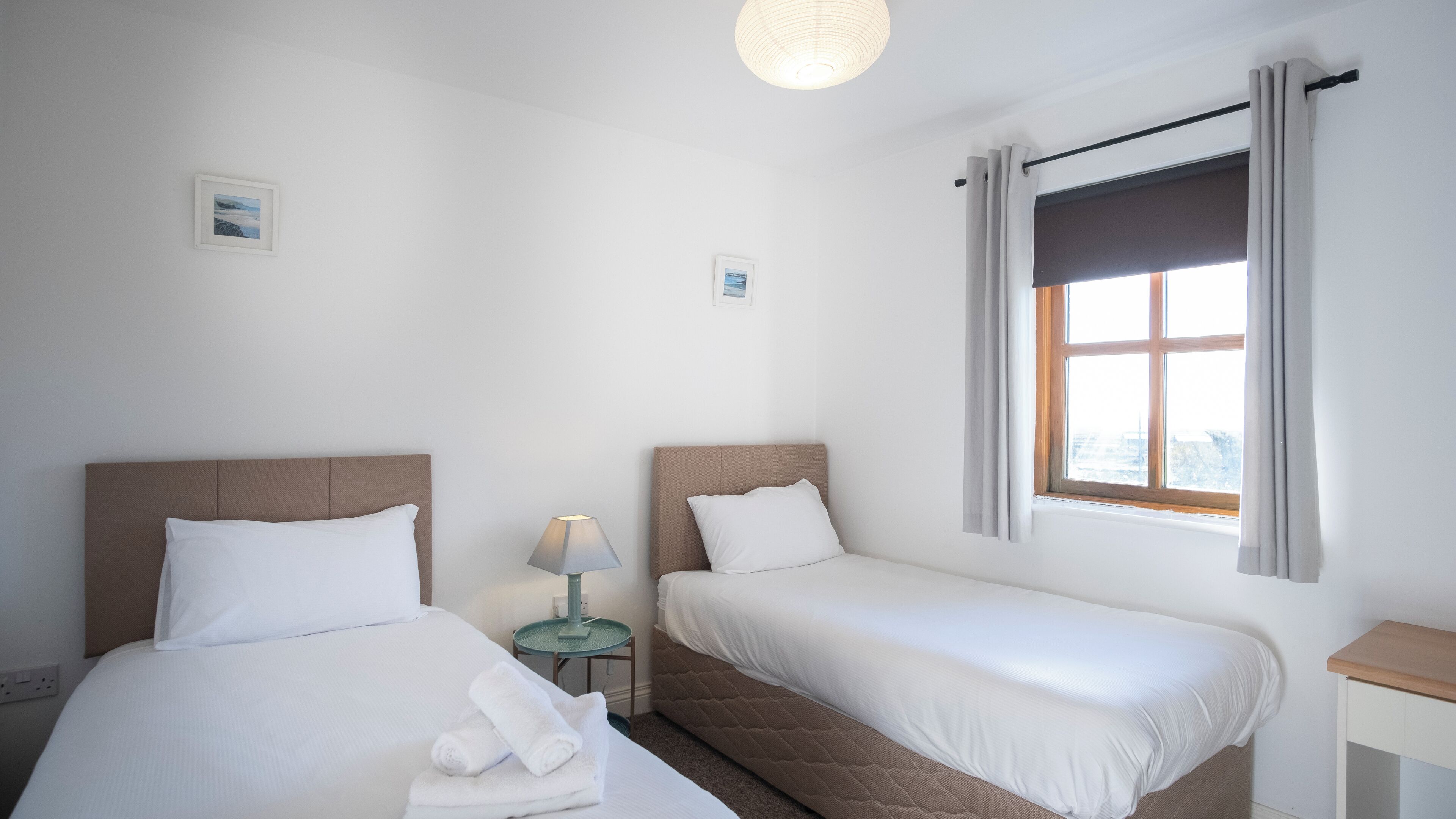 Habitación de hotel con dos camas individuales, una mesita de noche con lámpara, una ventana con cortinas y un aplique de pared.