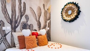 Une chambre avec un mur décoré sur le thème des cactus, un miroir décoratif et des coussins en forme de cœur.