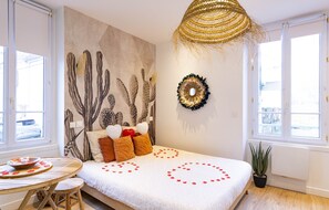Ein Schlafzimmer mit Bett, Wandgemälde, Pendelleuchte, Topfpflanze und Fenster.