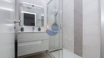 Un baño moderno con cabina de ducha de vidrio, tocador blanco con espejo y pared de ducha con azulejos.