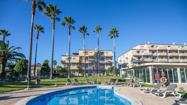 A swimming pool surrounded by palm trees and lounge chairs, with a multi-story building in the background. - 45 (Portimão)