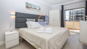 A modern bedroom with a grey headboard, white bedding, and a view of a balcony with buildings in the background. - 45 (Portimão)