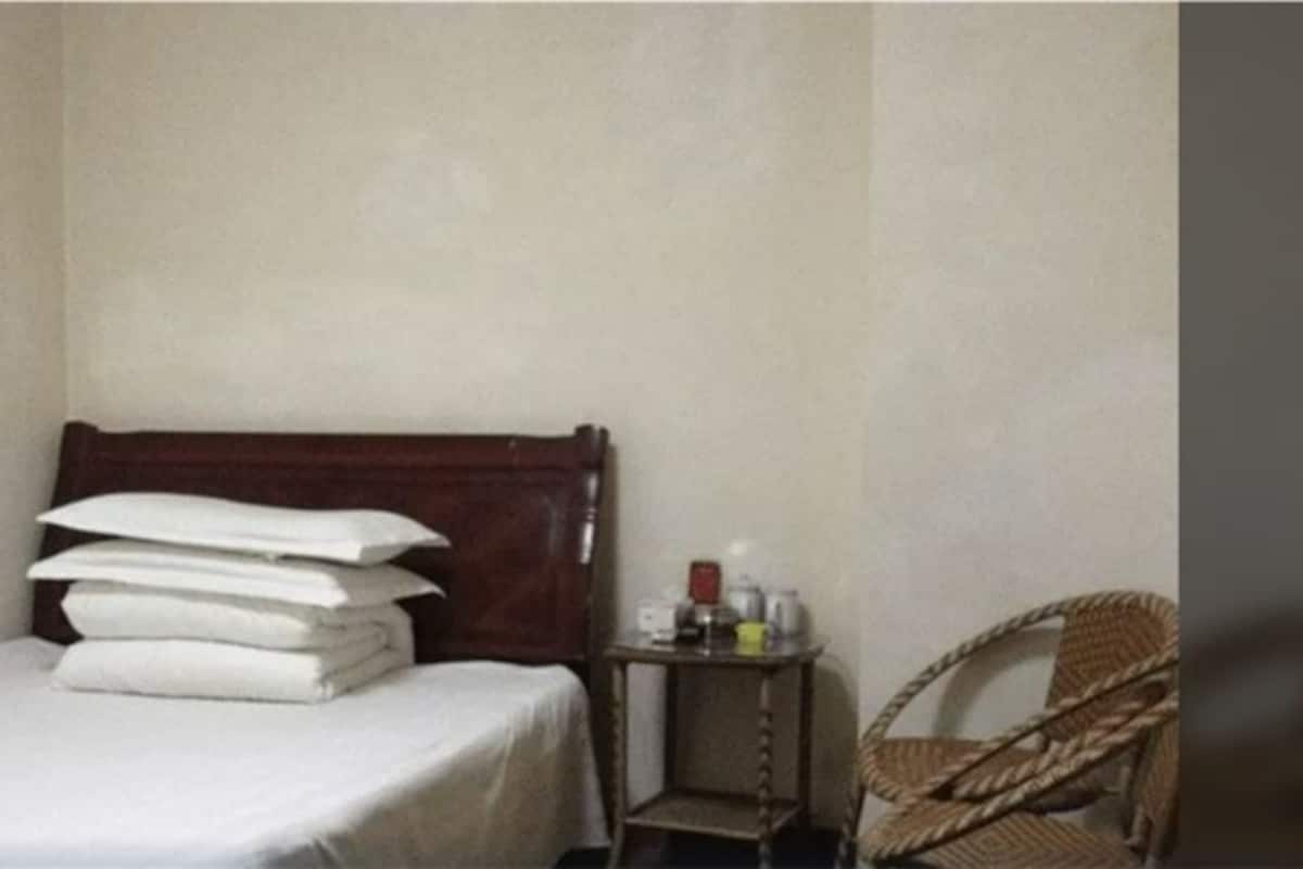 A single bed with white bedding, a small bedside table with a bottle, and a wicker chair.