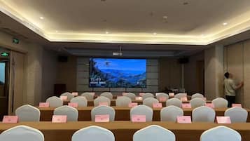 Un salón de conferencias con filas de sillas blancas, una pantalla grande al frente y un proyector.