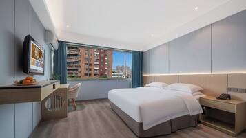 A modern hotel room with a bed, desk, chair, and a city view.