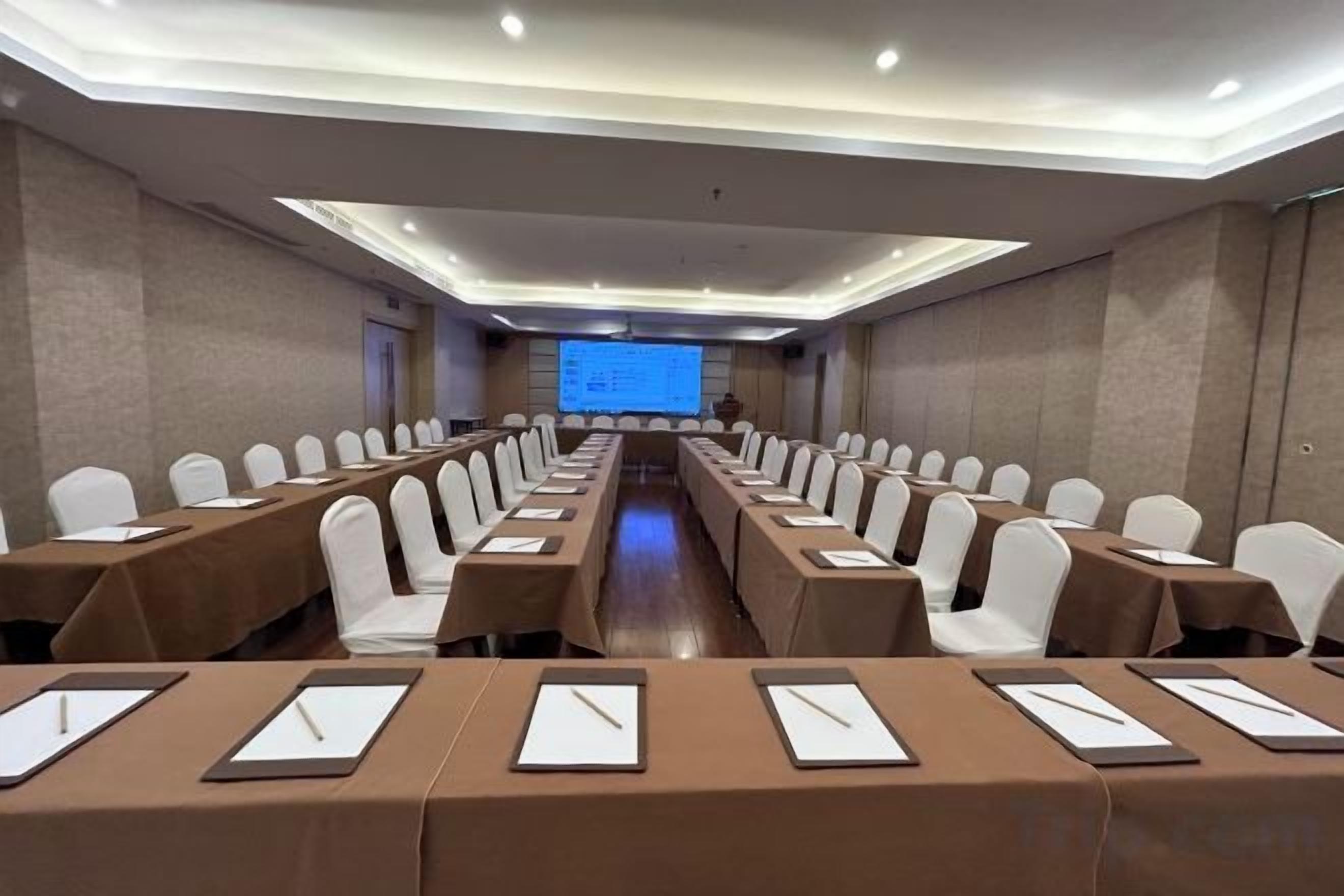 A conference room with rows of tables and chairs, a projector screen at the front, and a presentation visible on the screen.