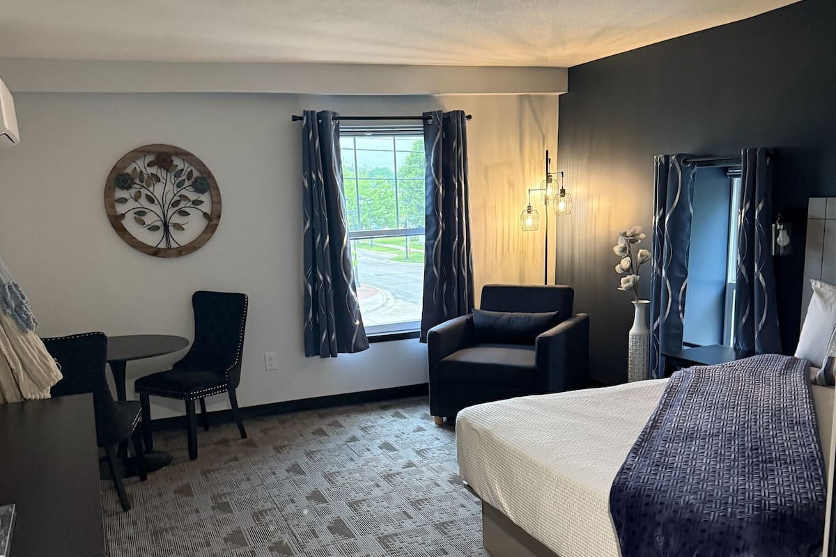 A hotel room with a bed, a desk, a chair, a sofa, and a large window with curtains.