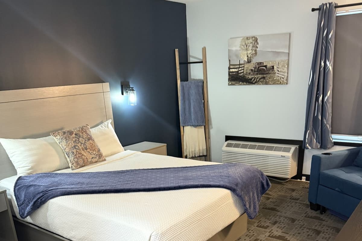 A neatly arranged hotel room with a bed, bedside tables, a blue chair, a standing heater, and a wall-mounted air conditioner.