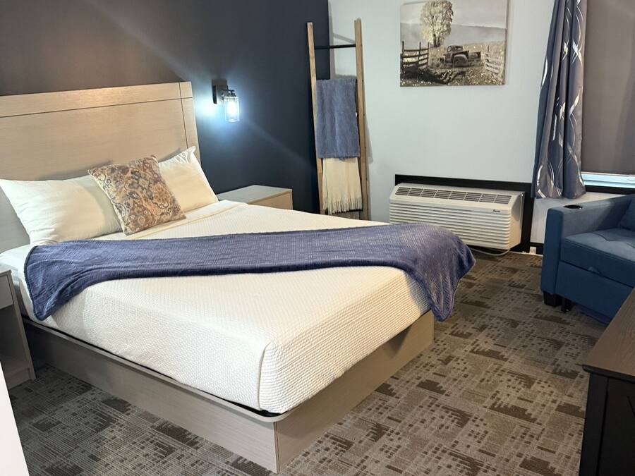 A neatly arranged hotel room with a bed, bedside tables, a blue chair, a standing heater, and a wall-mounted air conditioner.