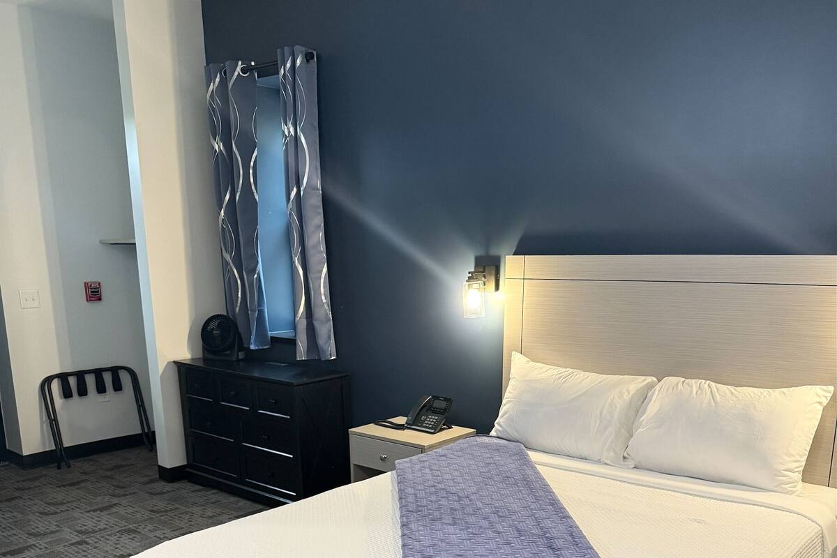 A hotel room with a bed, a bedside table with a phone, a wall-mounted light, a dark-colored headboard, a window with curtains, and a fire alarm on the wall.