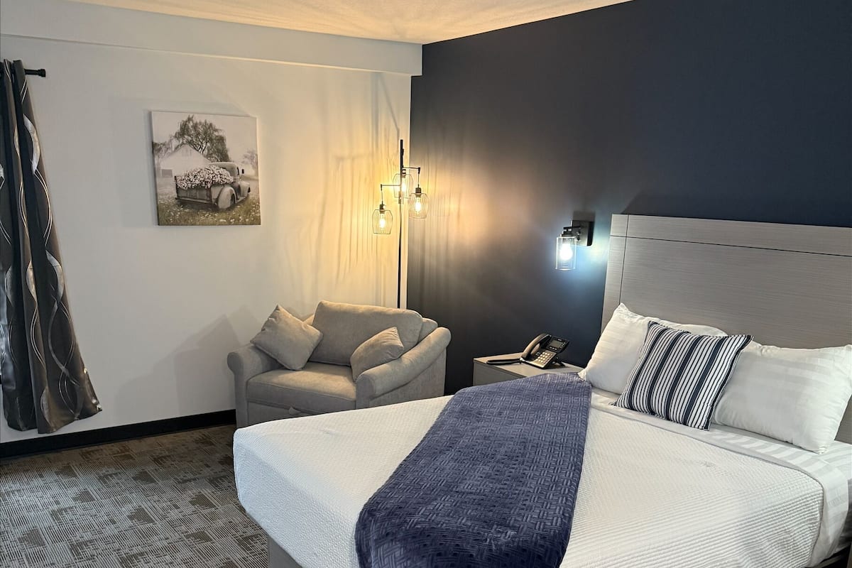 A hotel room with a large bed, a chair, a desk with a phone, and a wall-mounted lamp.
