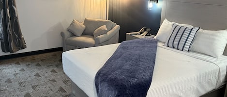 Hotellihuone, jossa on suuri sänky, tuoli, kirjoituspöytä puhelimineen ja seinään kiinnitetty lamppu.