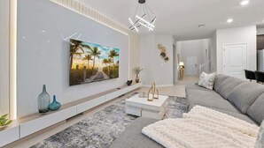 A modern living room with a large flat-screen TV mounted on the wall, a grey sofa, a white coffee table, and a patterned rug. - Super Homey (Clermont)
