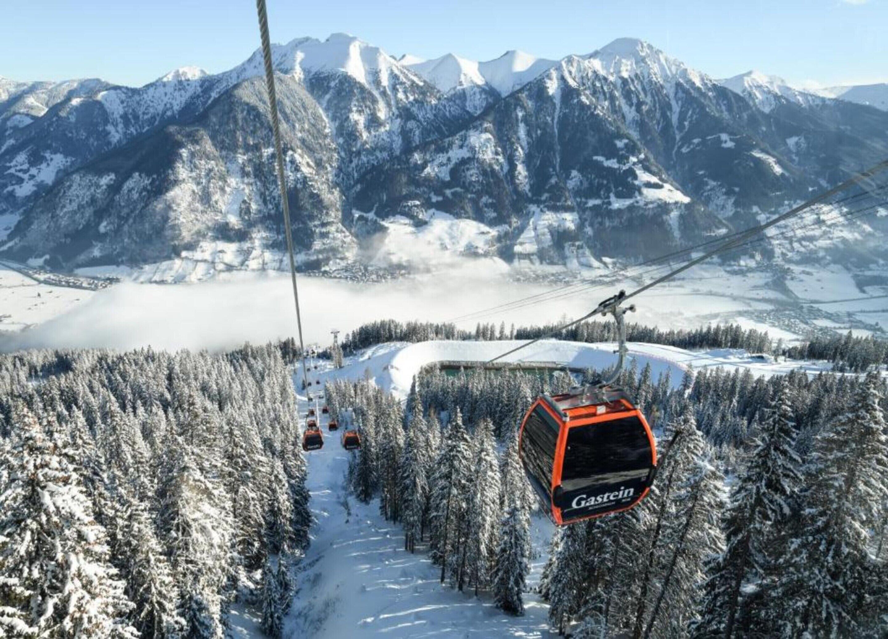 A cable car with the label "Gastein" moving along a snowy mountain path surrounded by dense forests and snow-covered peaks.