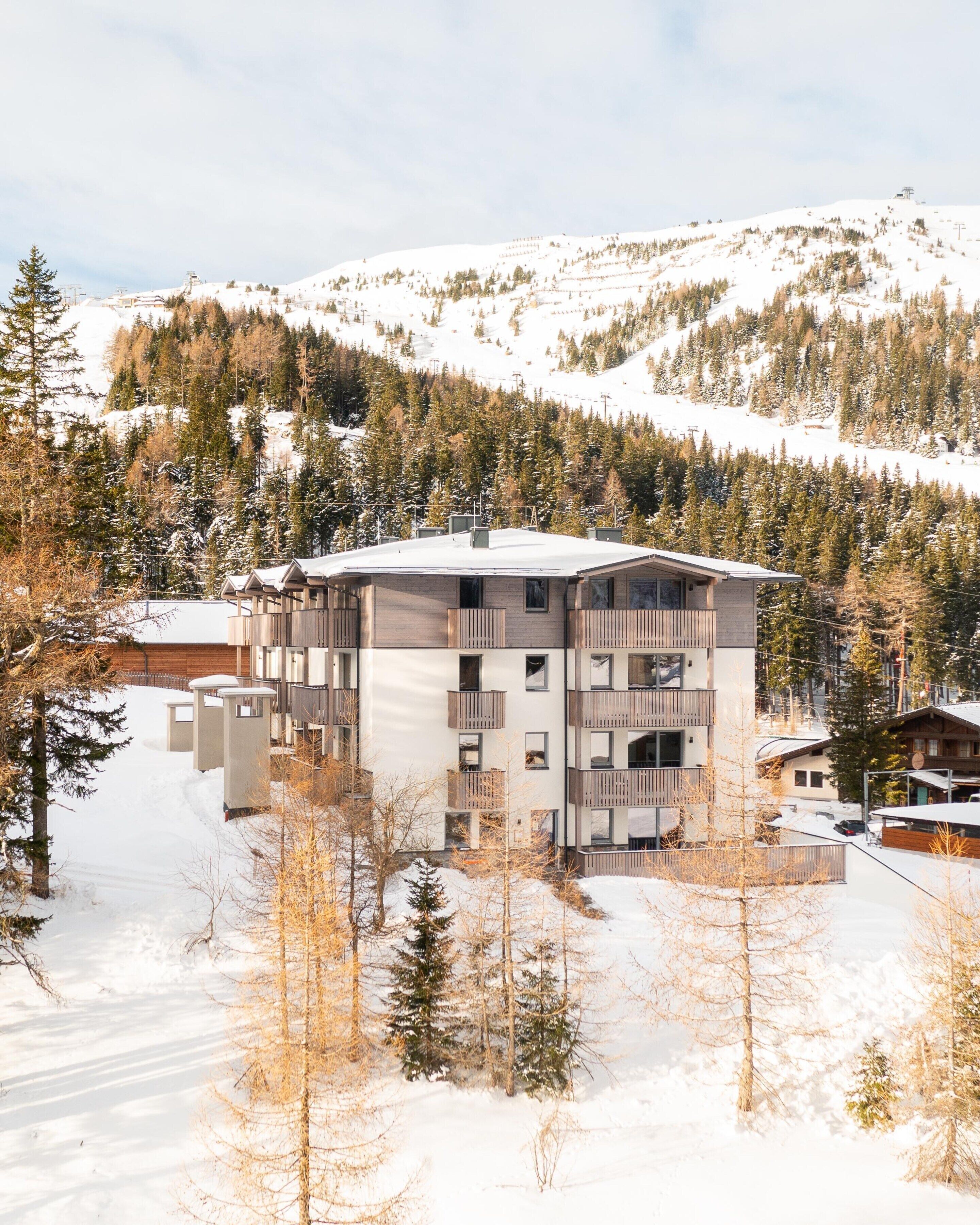 Un immeuble à plusieurs étages doté de balcons, entouré d’arbres couverts de neige et avec une chaîne de montagnes en arrière-plan.