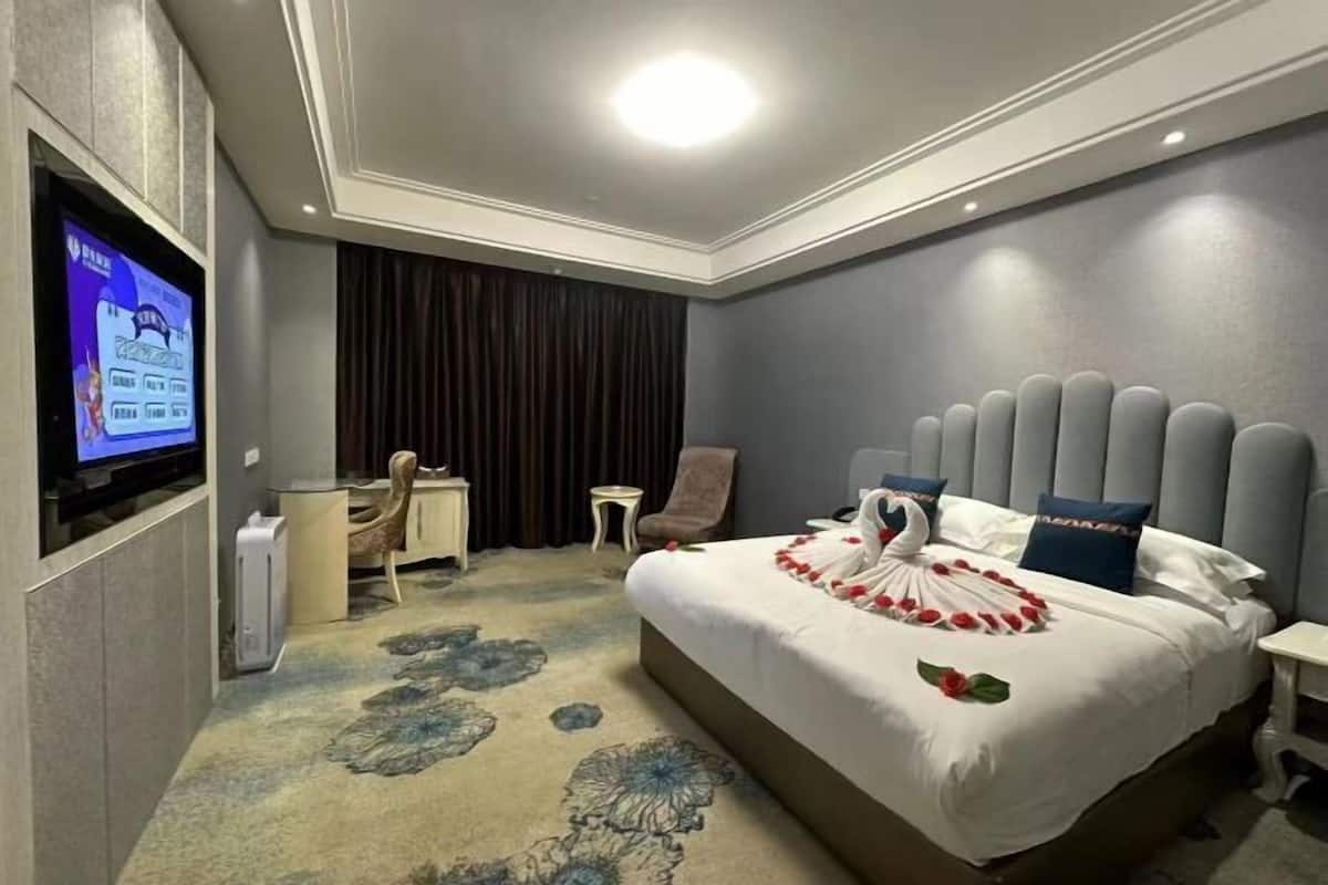 A hotel room with a large bed, a flat-screen TV mounted on the wall, a small desk with a chair, and a dark curtain.