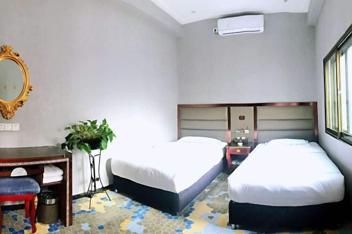A hotel room with two beds, a desk with a chair, a mirror, a potted plant, and an air conditioning unit.