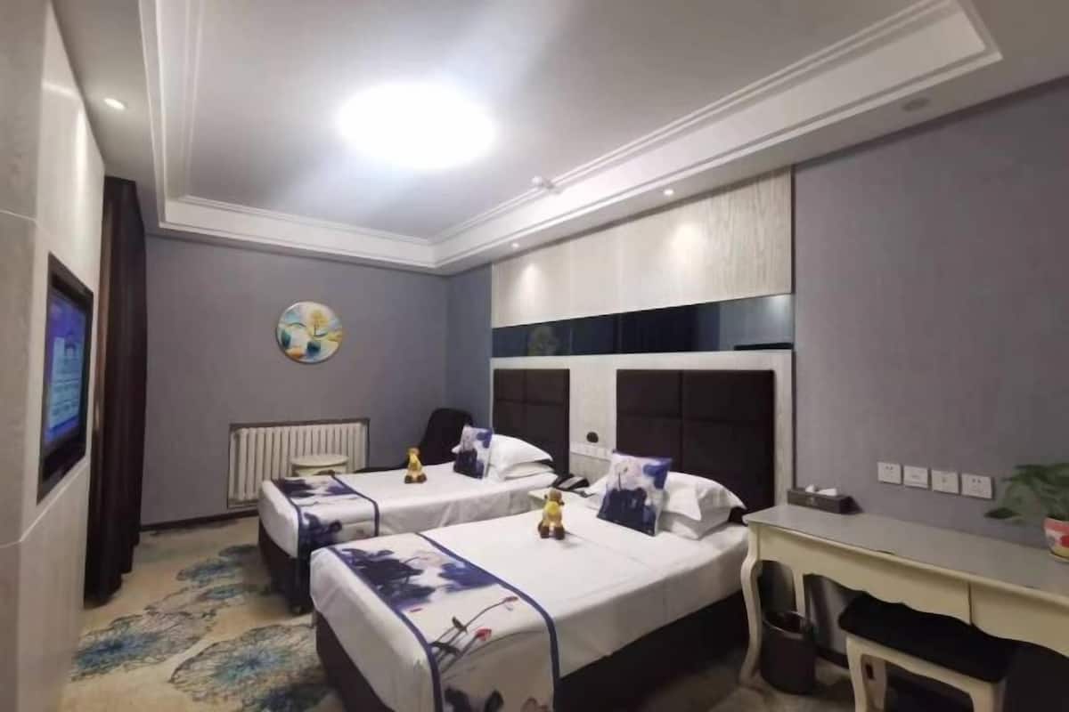 A hotel room with two beds, a desk, a radiator, a television, and a painting on the wall.