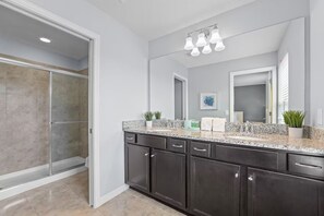 A modern bathroom with a glass shower enclosure, dark cabinetry, granite countertops, and a large mirror with a ceiling light fixture. - FL- 1844 GOBI DR (Kissimmee)