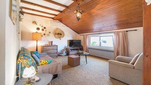 Un salon chaleureux avec un téléviseur, un canapé et un fauteuil, des poutres en bois au plafond et une fenêtre avec des rideaux.